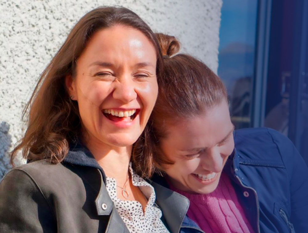 Two women laughing warmly together outdoors in natural sunlight.