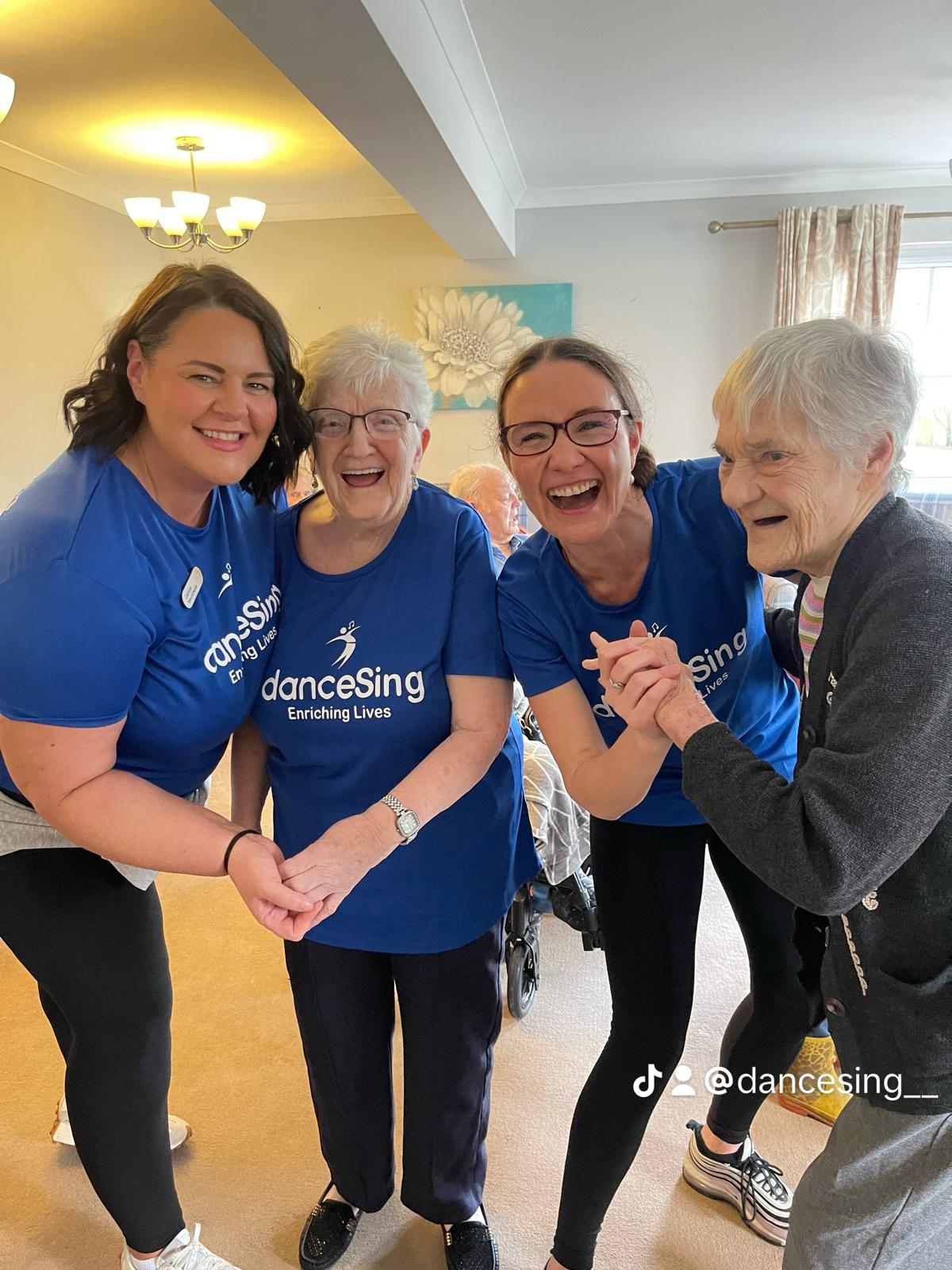 Group of women in blue shirts smiling and dancing with elderly ladies.