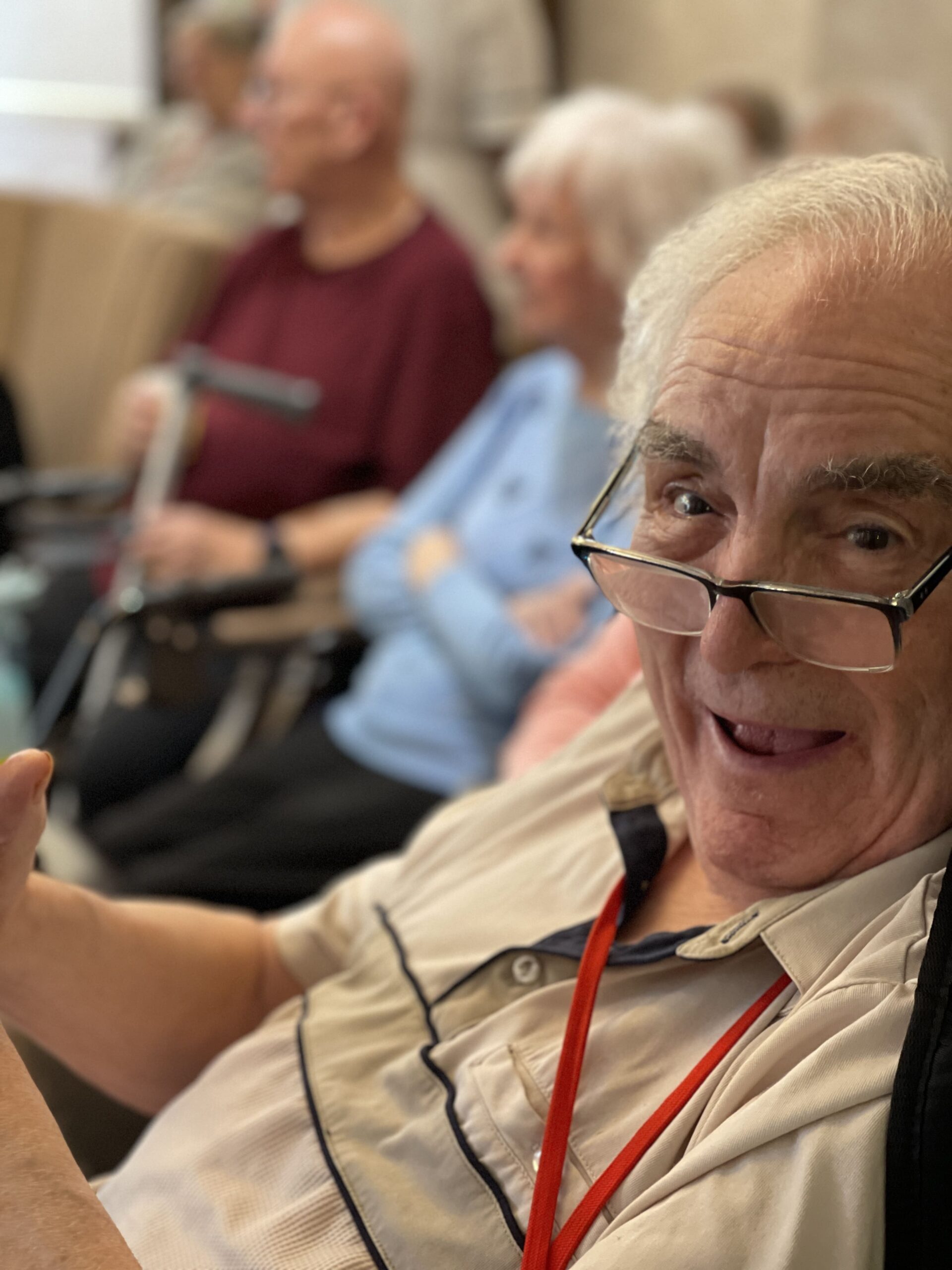 Elderly man wearing glasses smiling while sitting among other seniors.